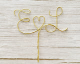 A personalized wire cake topper designed with the initials E and L with a heart shape between laying on a wood surface. 