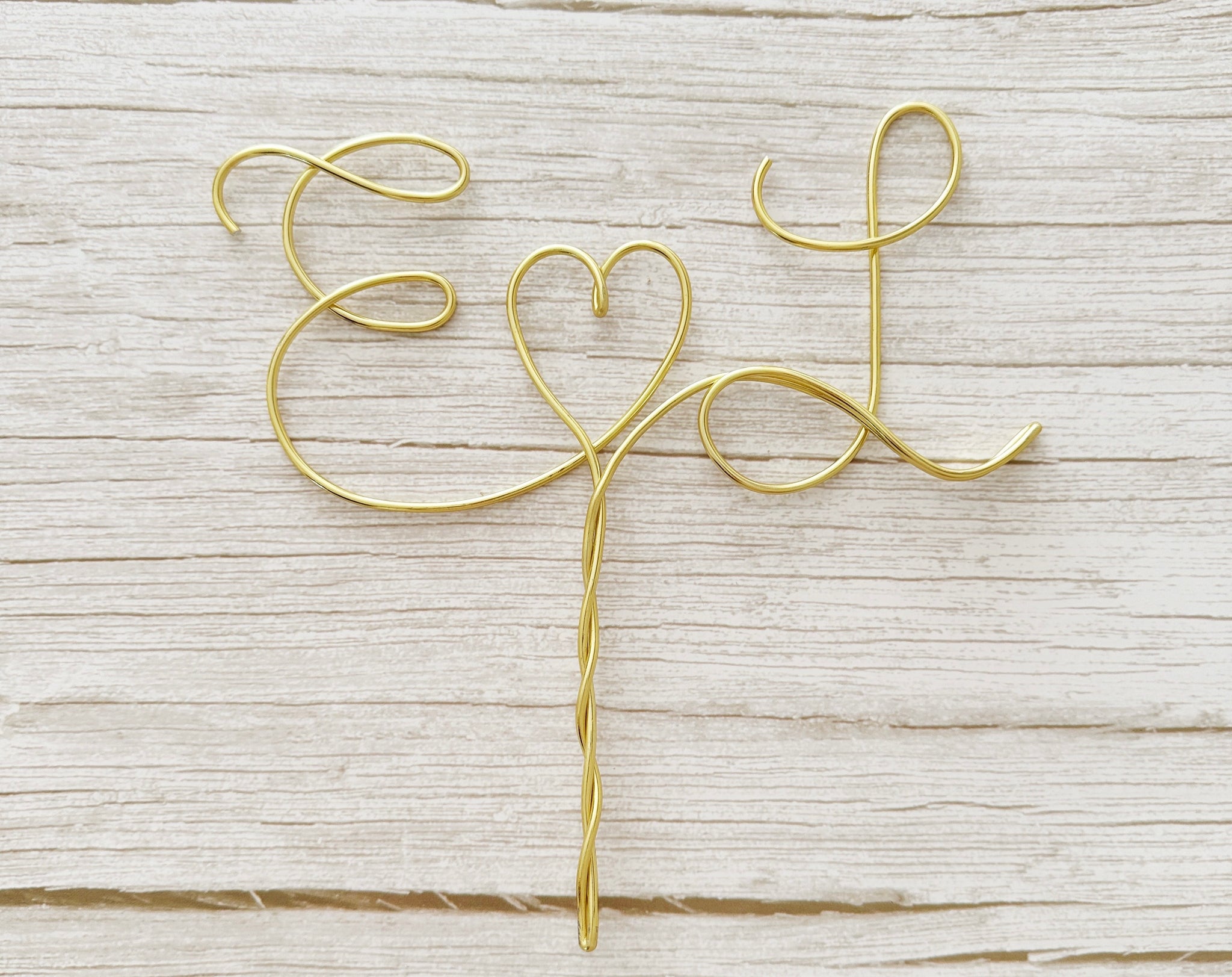 A personalized wire cake topper designed with the initials E and L with a heart shape between laying on a wood surface. 