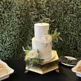 A wedding cake with a personalized wire cake topper designed with the initials E and L with a heart shape between. Cake adorned with a white rose and green leaves, set against a backdrop of green artificial foliage.