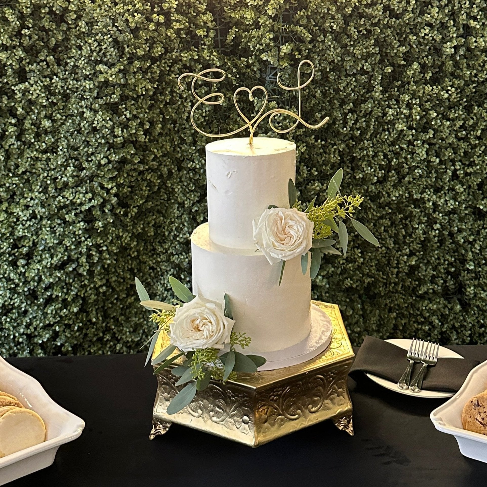 A wedding cake with a personalized wire cake topper designed with the initials E and L with a heart shape between. Cake adorned with a white rose and green leaves, set against a backdrop of green artificial foliage.