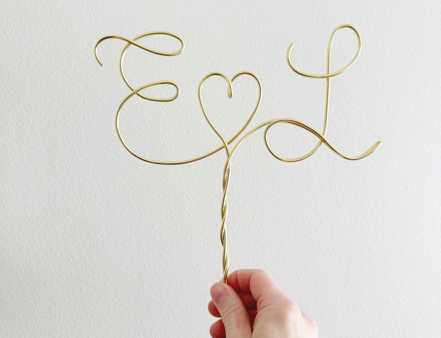 A personalized wire cake topper designed with the initials E and L with a heart shape between being help up in the air by a hand.