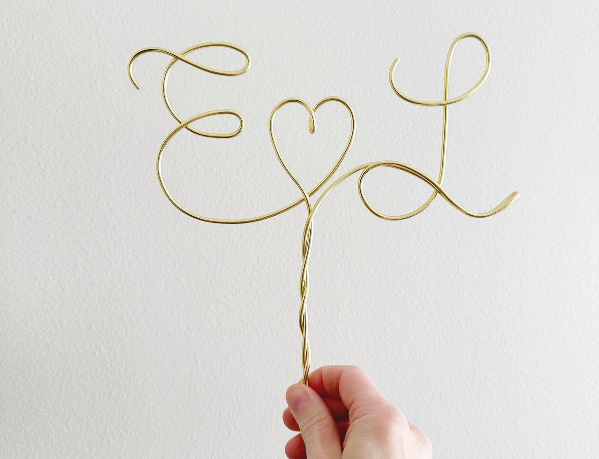 A personalized wire cake topper designed with the initials E and L with a heart shape between being help up in the air by a hand.