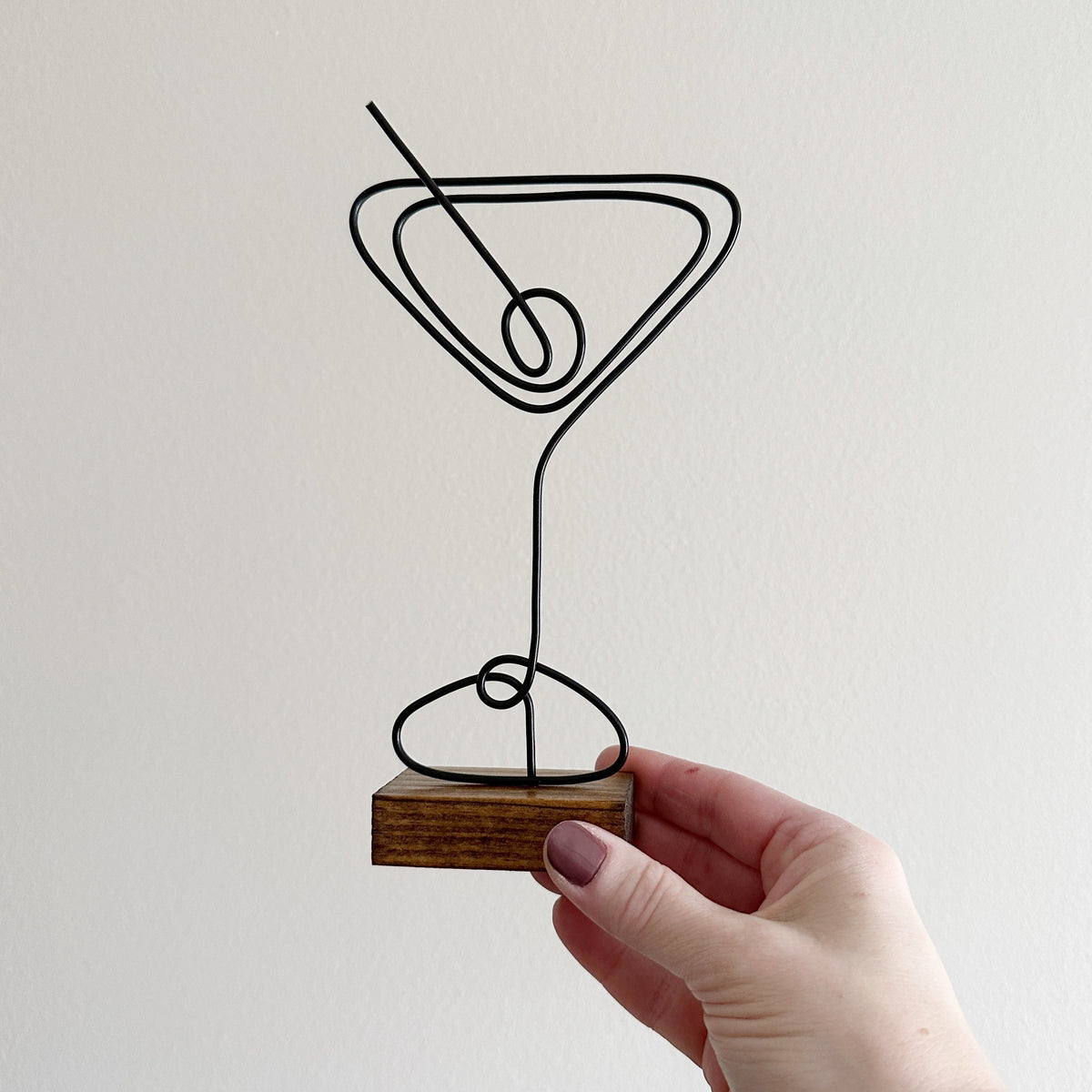 A hand holding a decorative wire martini glass sign mounted on a wooden base.