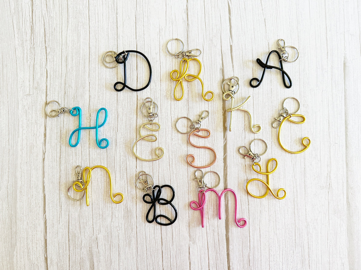 Twisted wire alphabet initial charms in various colors (black, gold, blue, pink, silver, rose gold) arranged on a white wood background.