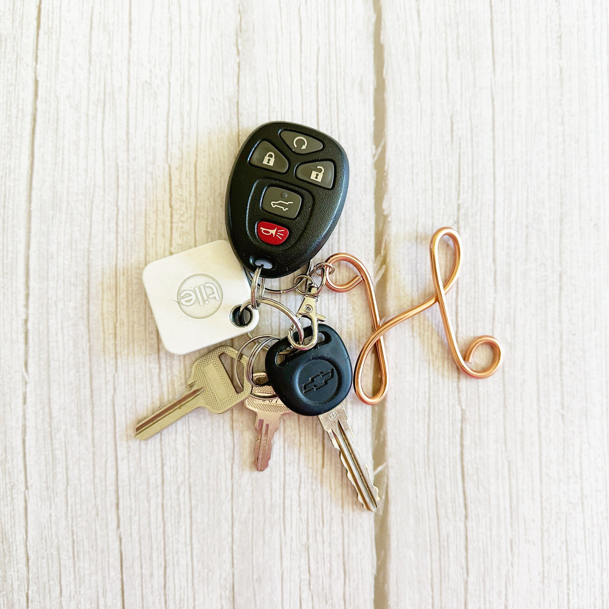 A set of car keys and key fobs arranged on a light wood surface. The set includes a black remote key fob, a white square key tag with a circular logo, several metal keys, and a copper-colored decorative keychain loop.