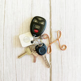 A set of car keys and key fobs arranged on a light wood surface. The set includes a black remote key fob, a white square key tag with a circular logo, several metal keys, and a copper-colored decorative keychain loop.