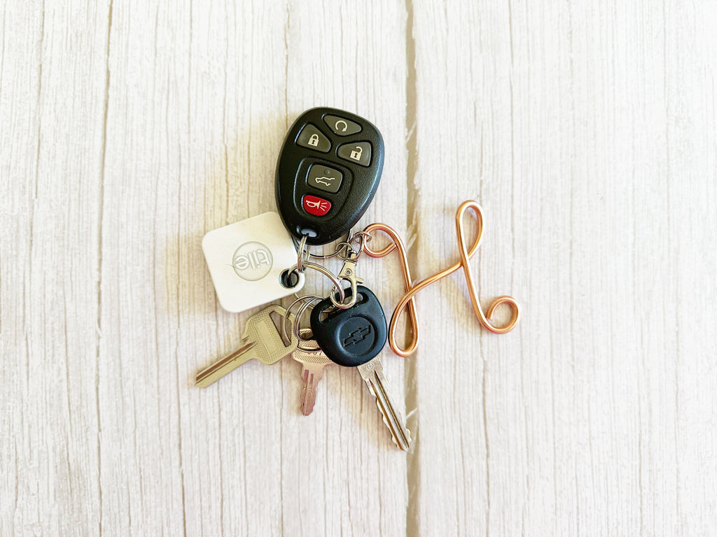 A set of car keys and key fobs arranged on a light wood surface. The set includes a black remote key fob, a white square key tag with a circular logo, several metal keys, and a copper-colored decorative keychain loop.