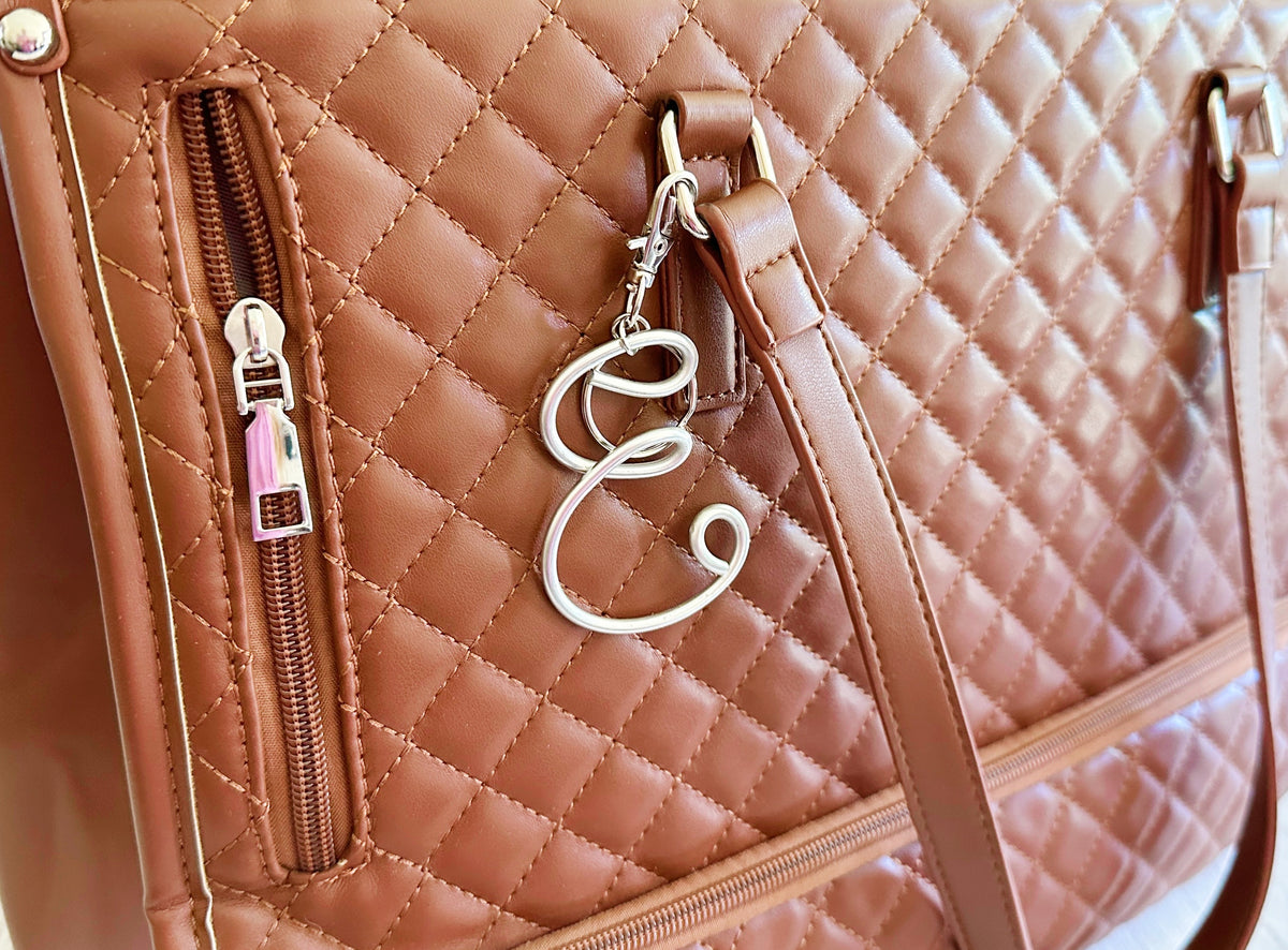 Close-up of a quilted tan leather handbag with a diagonal zip pocket, a silver-tone monogram keychain letter 'E', and a matching shoulder strap.