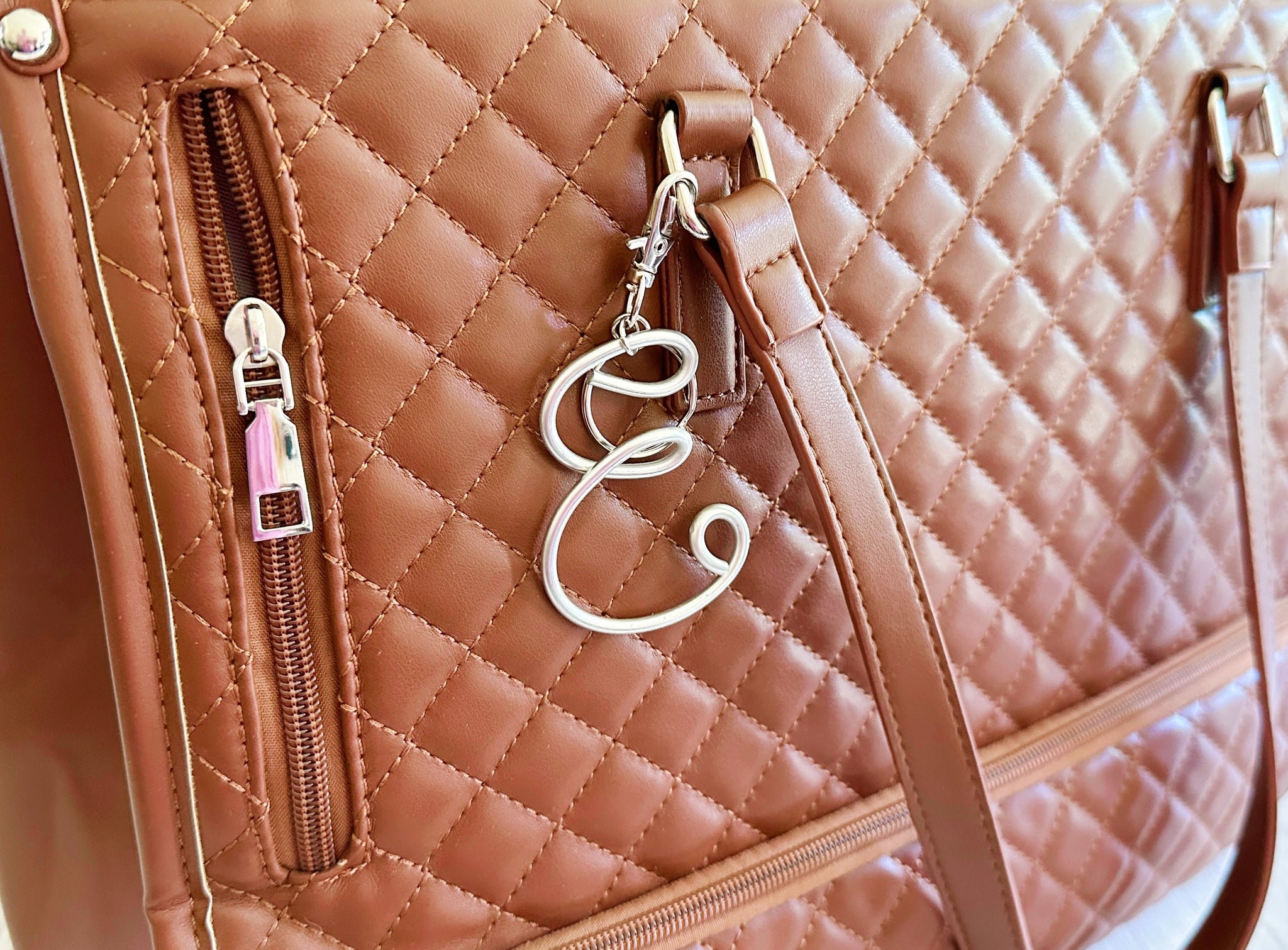 Close-up of a quilted tan leather handbag with a diagonal zip pocket, a silver-tone monogram keychain letter 'E', and a matching shoulder strap.