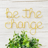 Gold wire art with 'be the change' on a light wooden background with green leaves.