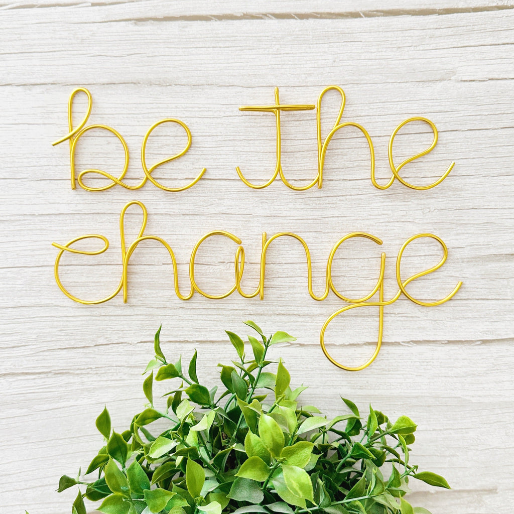 Gold wire art with 'be the change' on a light wooden background with green leaves.