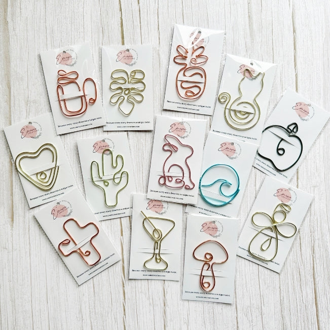 An assortment of colorful wire art clips/bookmarks shaped like fruits, cactus, and abstract designs, each mounted on a white backing card with a pink logo, arranged on a light wood background.
