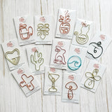 An assortment of colorful wire art clips/bookmarks shaped like fruits, cactus, and abstract designs, each mounted on a white backing card with a pink logo, arranged on a light wood background.