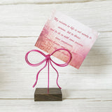 Card inserted into a wire pink bow photo holder on a wooden stand against a light wooden background