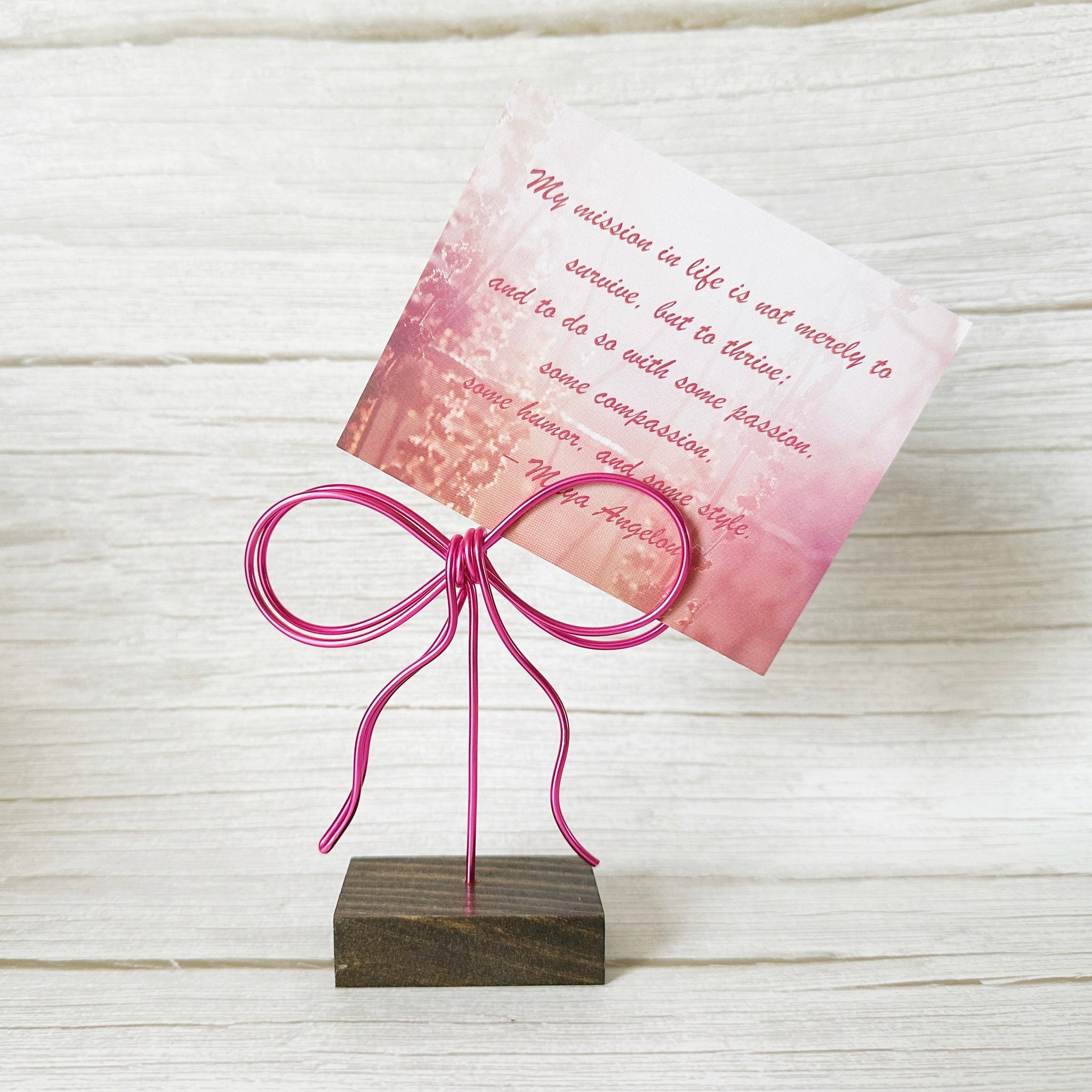 Card inserted into a wire pink bow photo holder on a wooden stand against a light wooden background