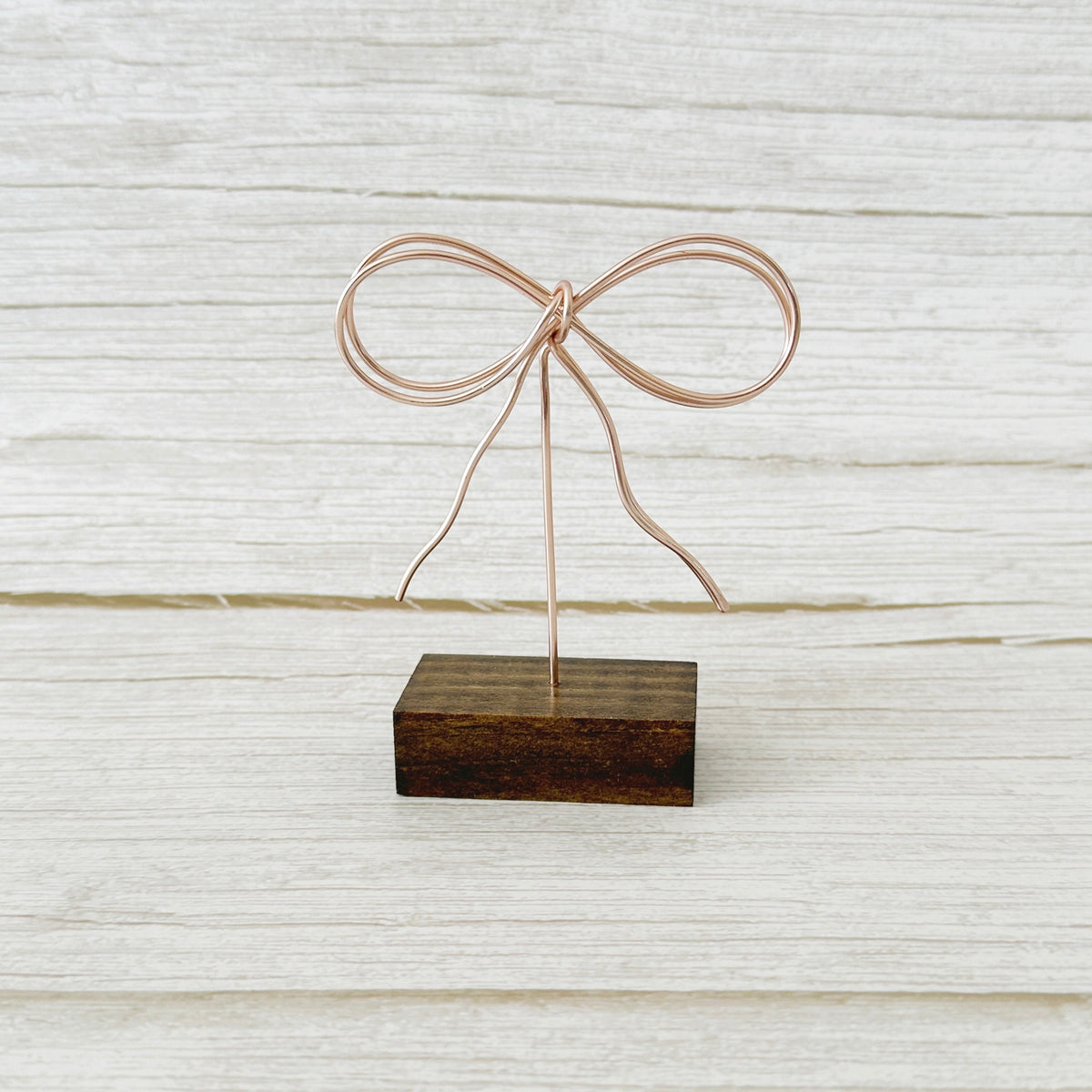 A bow-shaped wire art piece mounted on a wooden base, displayed against a wooden background.
