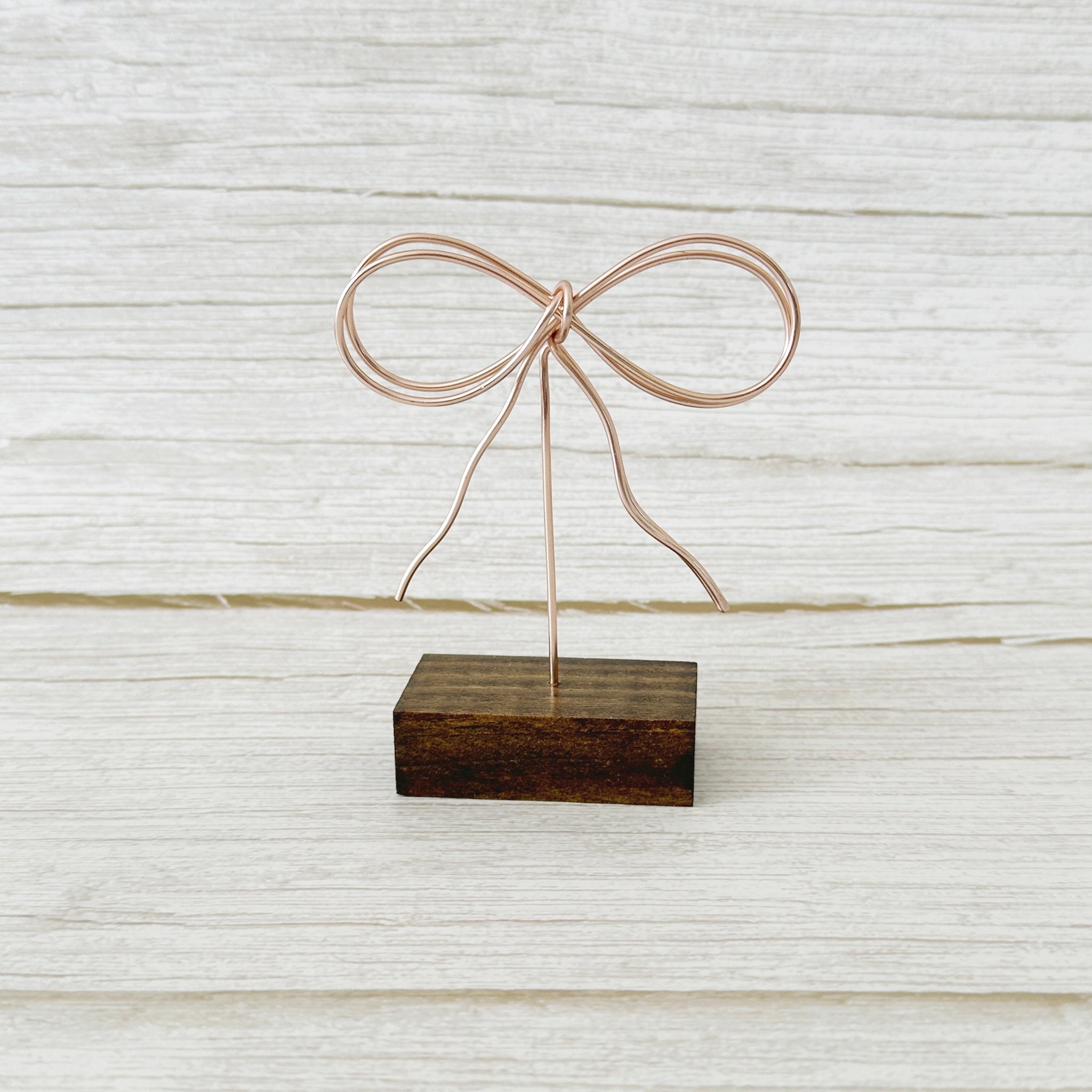 A bow-shaped wire art piece mounted on a wooden base, displayed against a wooden background.