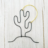 Handmade wire art of a black cactus and gold sun, styled on a light wood background—minimalist desert wall decor.