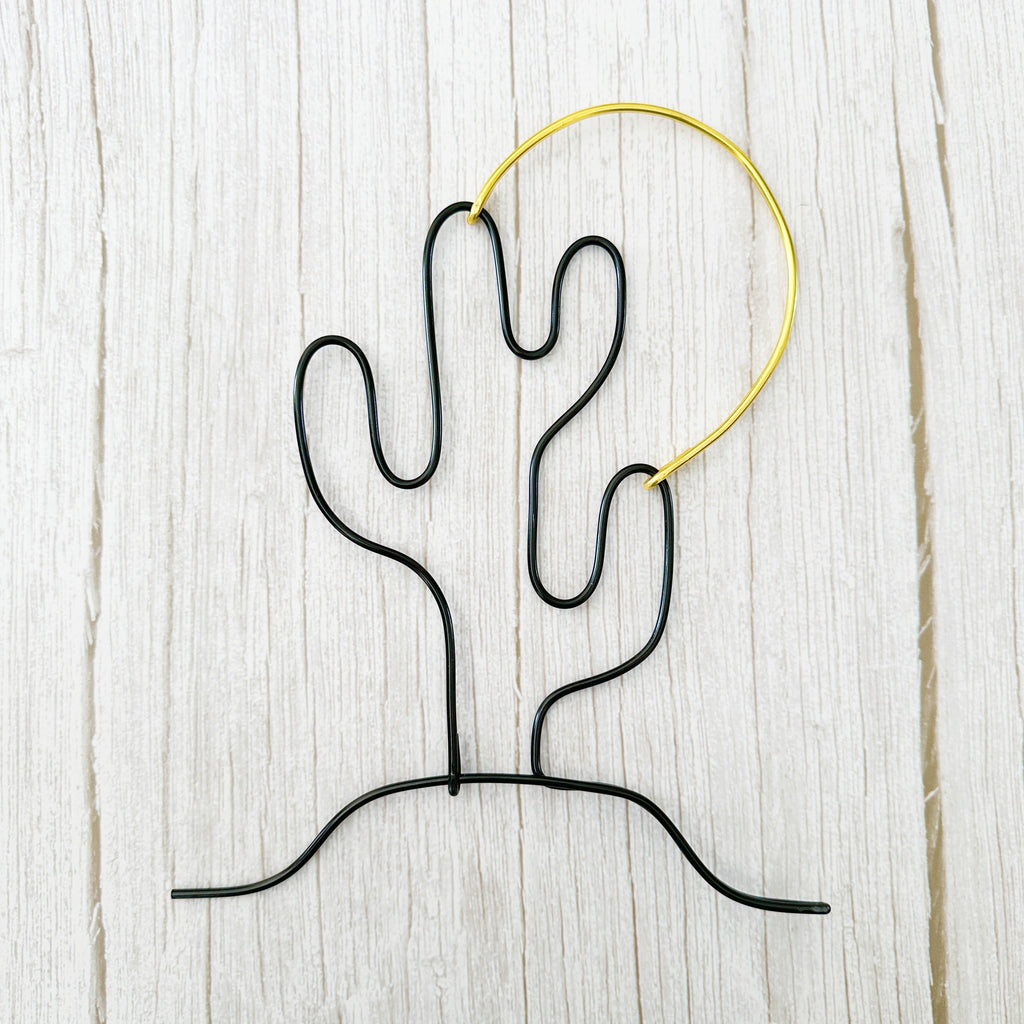 Handmade wire art of a black cactus and gold sun, styled on a light wood background—minimalist desert wall decor.