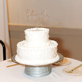 A wedding cake with a personalized wire cake topper designed with the initials K and J with a heart shape between. Cake is sitting on a metal cake stand on top of a table with plates and forks.
