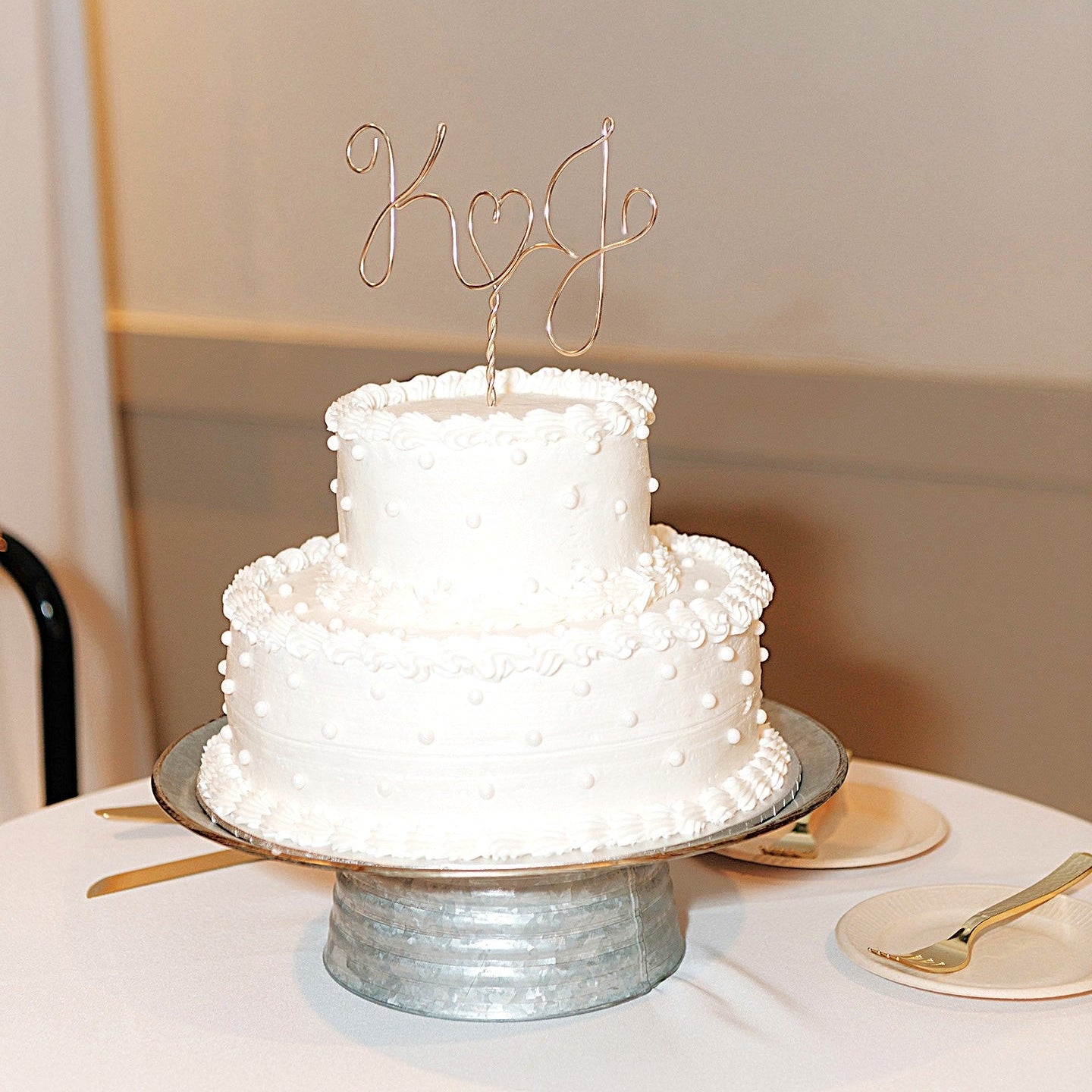 A wedding cake with a personalized wire cake topper designed with the initials K and J with a heart shape between. Cake is sitting on a metal cake stand on top of a table with plates and forks.