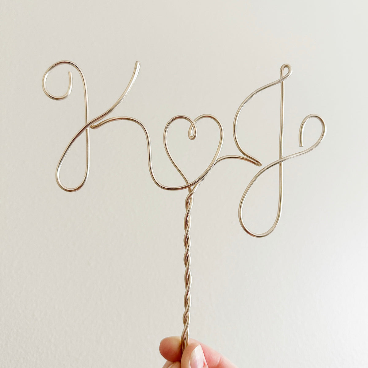 A personalized wire cake topper designed with the initials K and J with a heart shape between. Being held up in the air with two fingers.