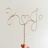 A personalized wire cake topper designed with the initials K and J with a heart shape between. Being held up in the air with two fingers.
