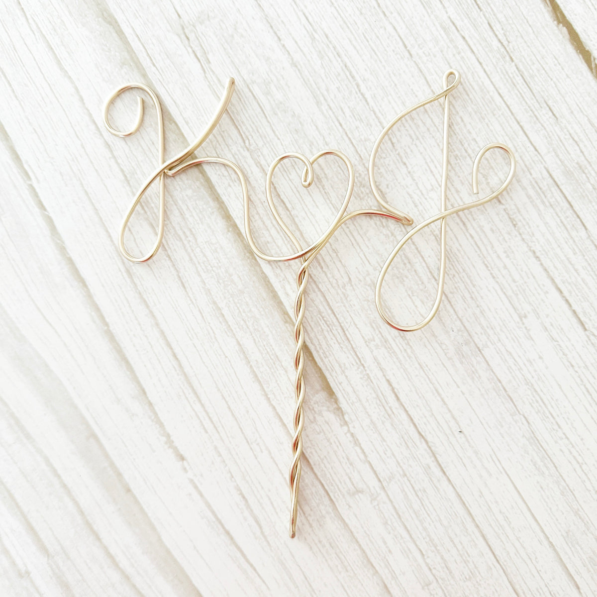 A personalized wire cake topper designed with the initials K and J with a heart shape between laying on a wood surface. 