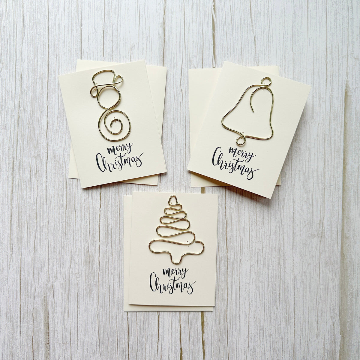Three Christmas-themed cards with handcrafted wire art designs of a snowman, bell and Christmas tree on a wooden surface.