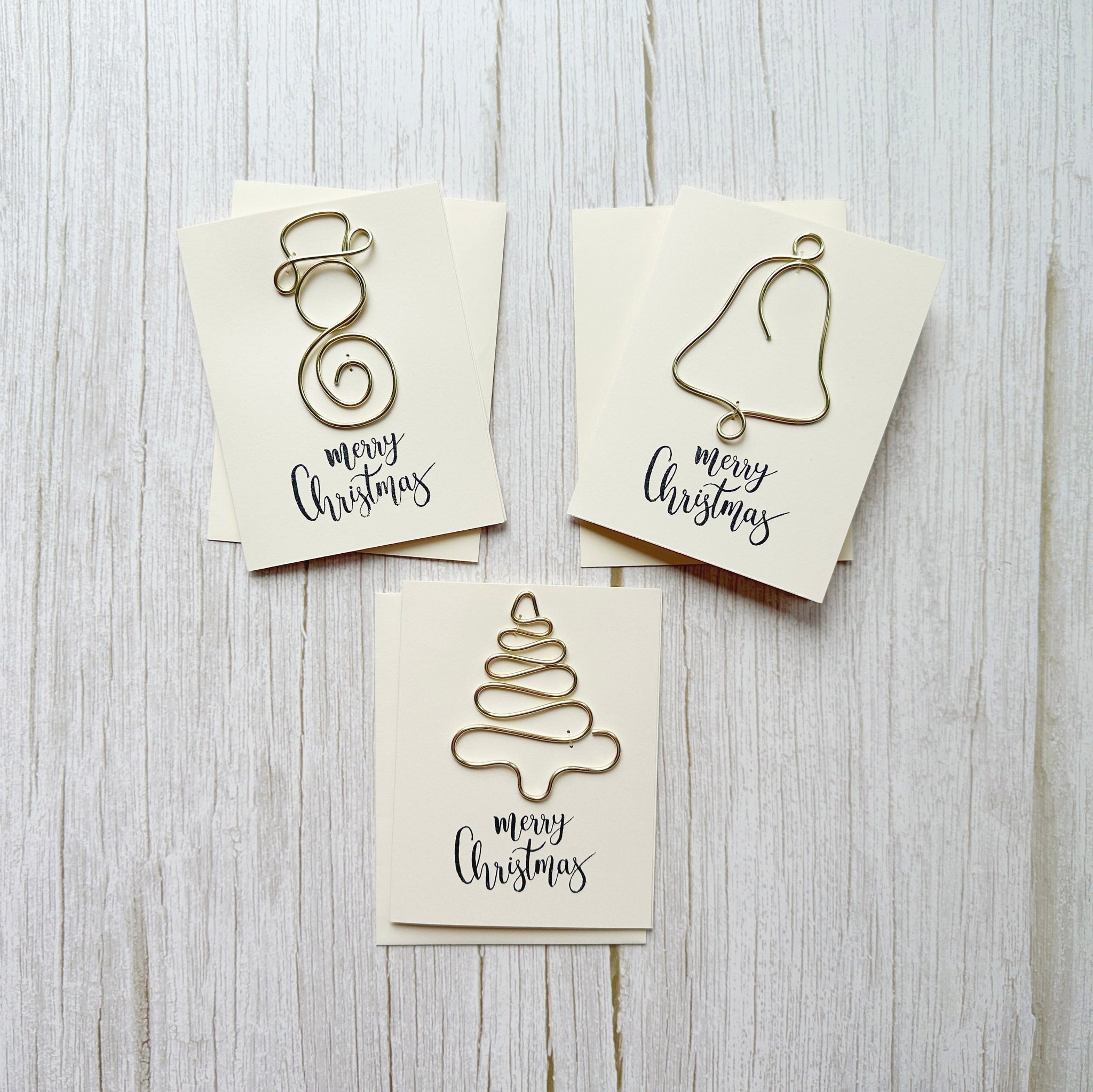Three Christmas-themed cards with handcrafted wire art designs of a snowman, bell and Christmas tree on a wooden surface.