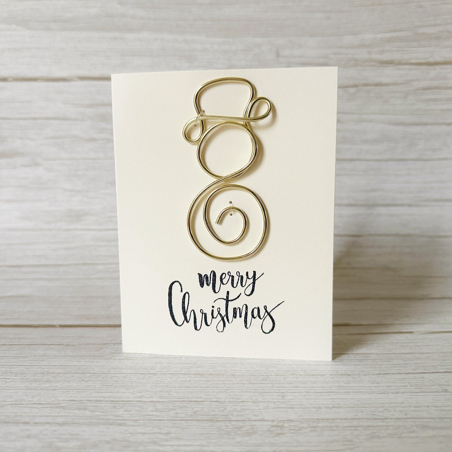 Gold wire Christmas decoration on a white card with 'Merry Christmas' text, placed on a wooden surface.