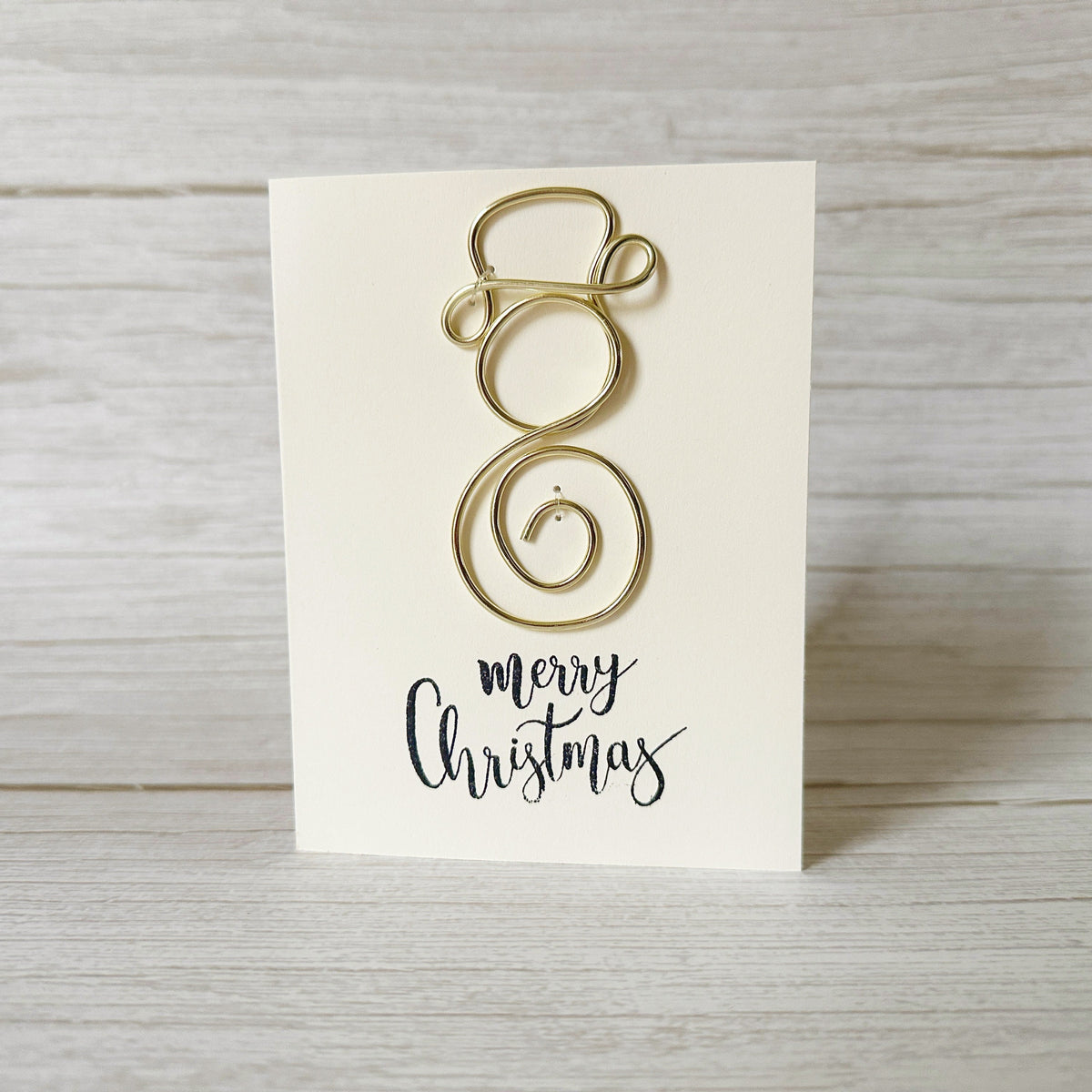 Gold wire Christmas decoration on a white card with 'Merry Christmas' text, placed on a wooden surface.