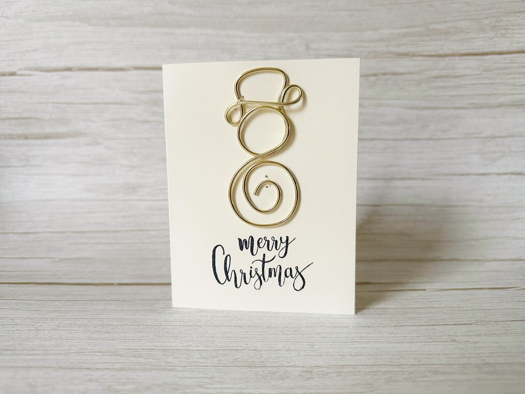 Gold wire Christmas decoration on a white card with 'Merry Christmas' text, placed on a wooden surface.