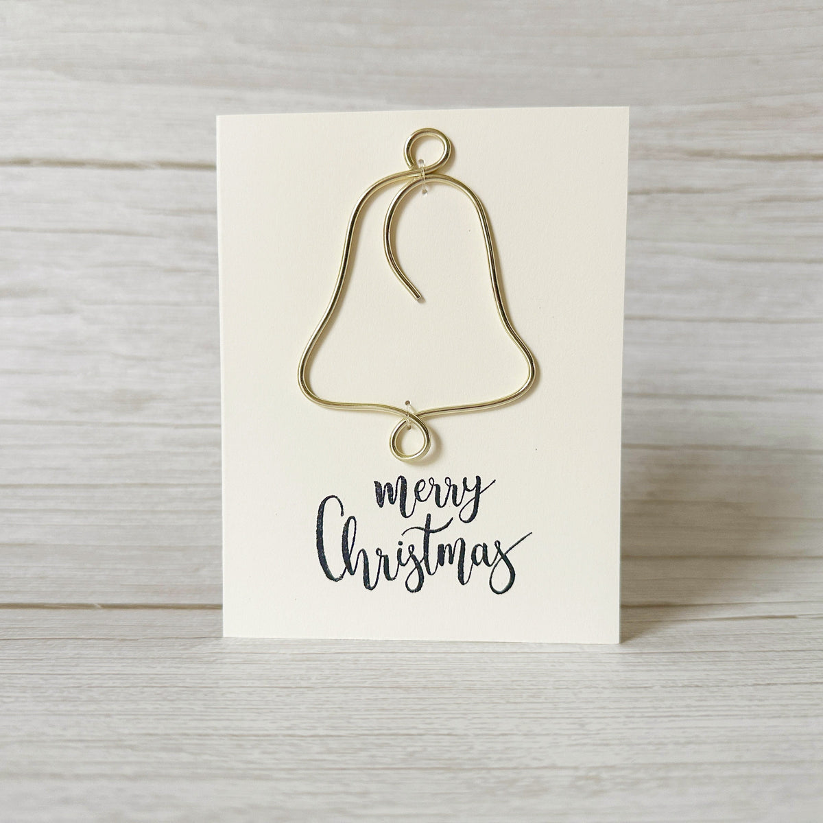 Gold Christmas bell ornament  handcrafted from wire on an ivory card with 'merry Christmas' text, against a light wooden background.