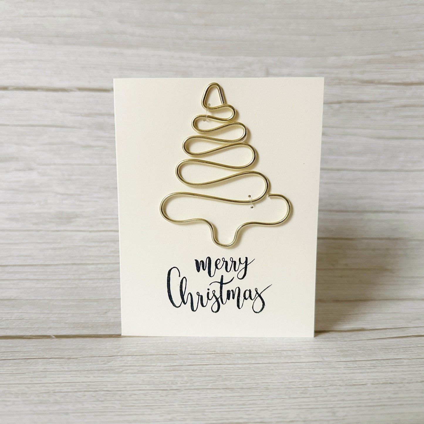 Gold Christmas tree-shaped ornament made from wire on a white card with 'merry Christmas' text, placed on a wooden surface.