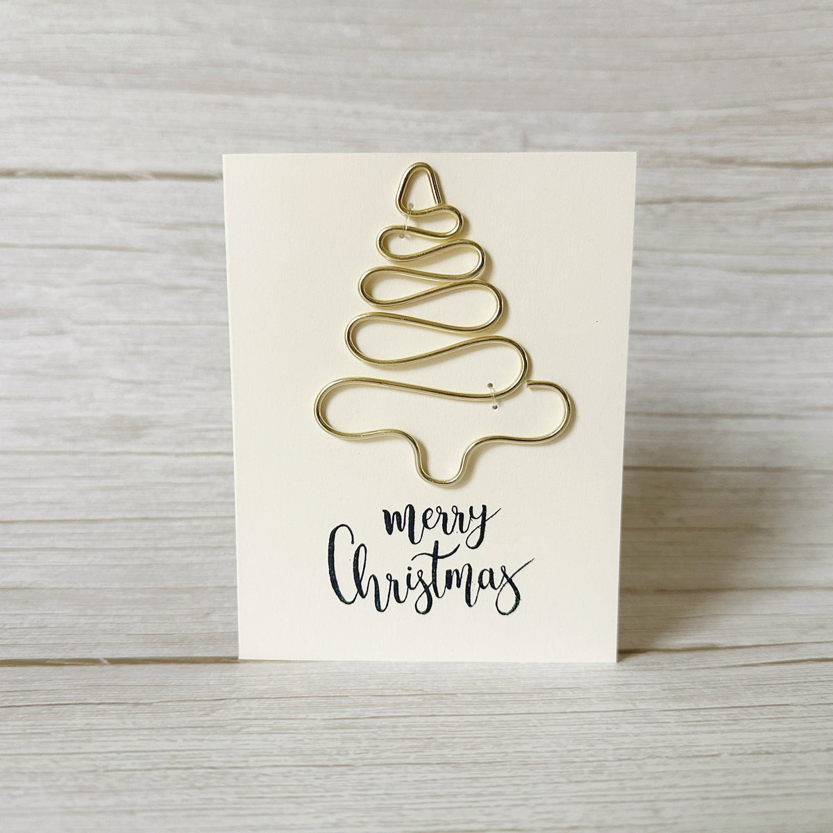 Gold Christmas tree-shaped ornament made from wire on a white card with 'merry Christmas' text, placed on a wooden surface.