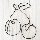 Minimalist cherry wall art made from black wire, shaped into two cherries with a leaf, styled on a light wood background.