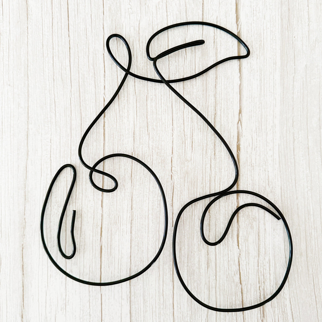 Minimalist cherry wall art made from black wire, shaped into two cherries with a leaf, styled on a light wood background.