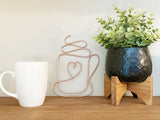 White mug, decorative coffee mug wire heart sculpture, and potted plant on a wooden surface with a white wall background.