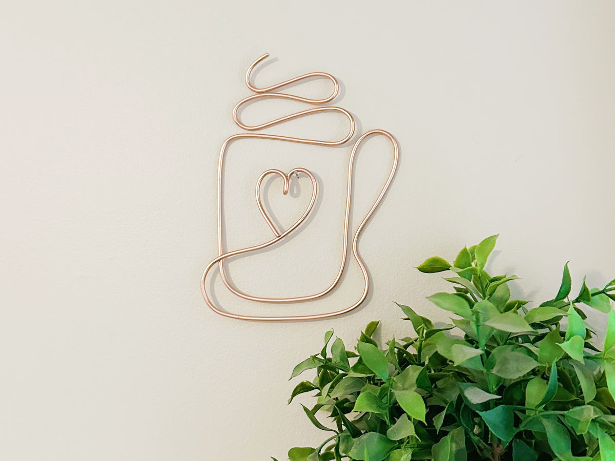 Decorative wire coffee mug wall art with a heart design on a light background and hanging from  a nail. A green faux plant is below.