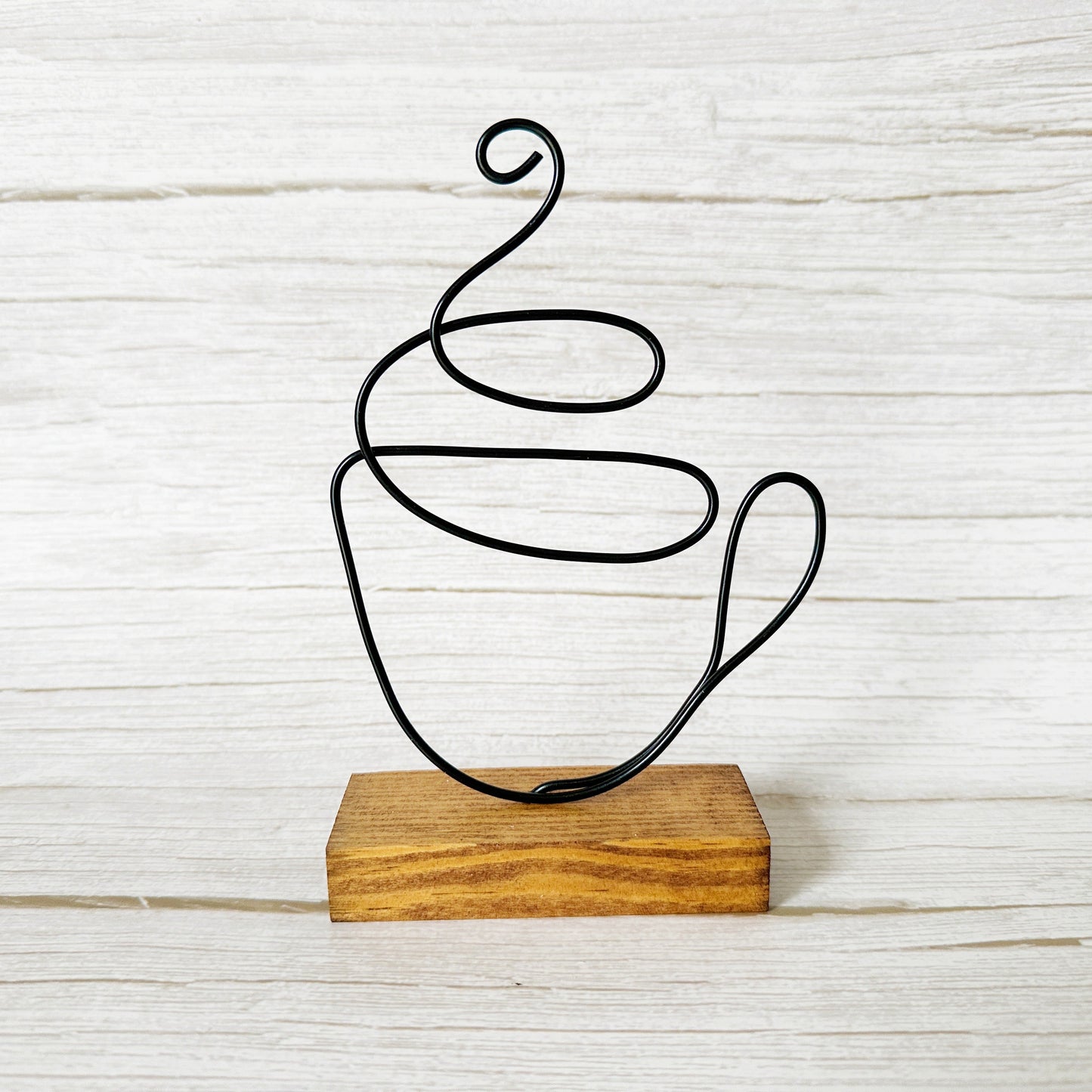 Handcrafted wire coffee cup sculpture in black wire, mounted on a stained wood base, styled against a white rustic background.