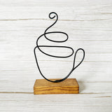 Handcrafted wire coffee cup sculpture in black wire, mounted on a stained wood base, styled against a white rustic background.