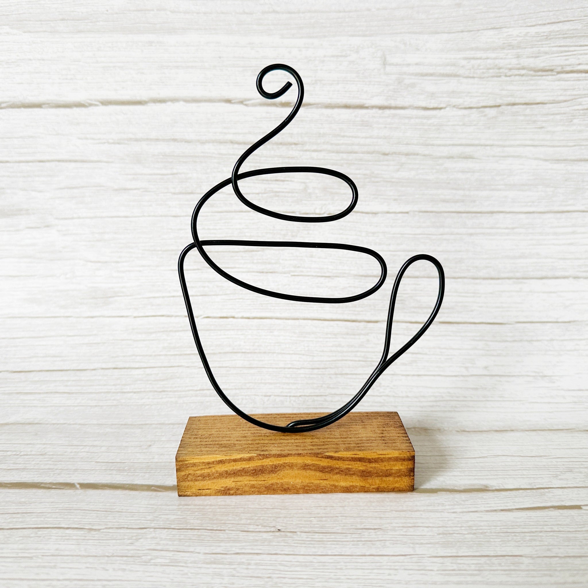 Handcrafted wire coffee cup sculpture in black wire, mounted on a stained wood base, styled against a white rustic background.