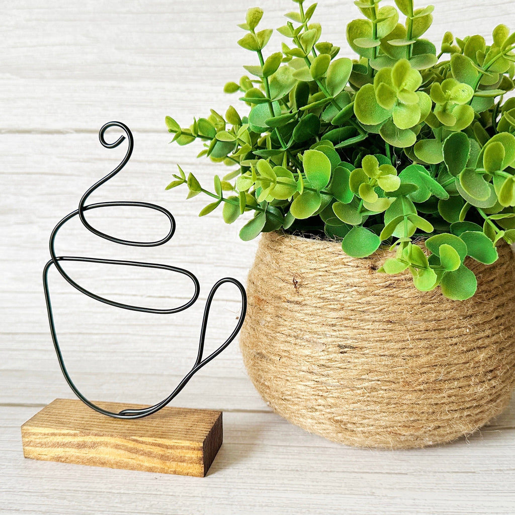 Decorative wire coffee mug sculpture and woven pot with green plant on a light wooden surface