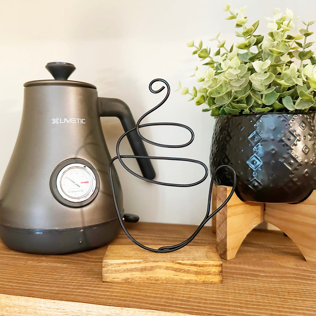 Wire coffee mug sculpture on a shelf between a silver electric kettle and a faux plant.
