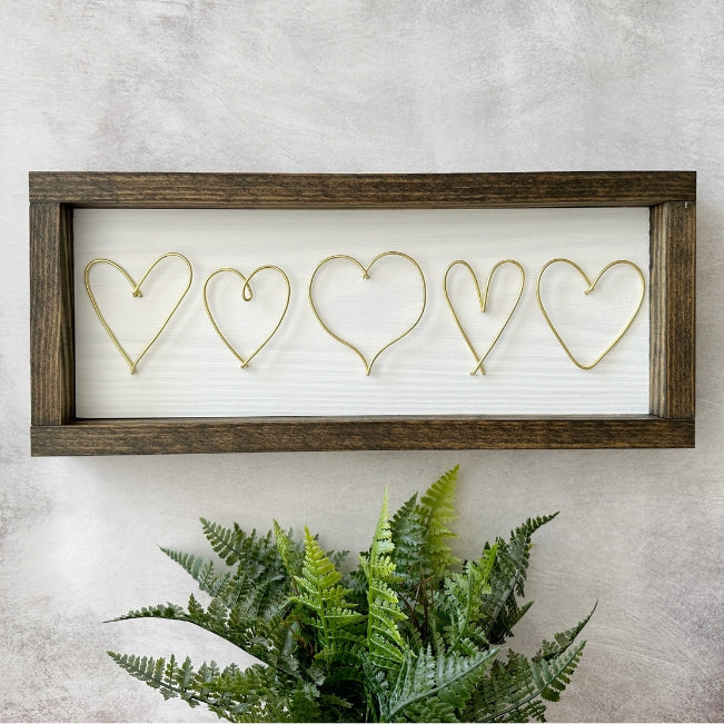 A rectangular framed wall decor piece with five wire heart outlines on a white background, mounted on a textured light gray wall above a green fern plant.