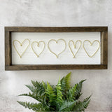 A rectangular framed wall decor piece with five wire heart outlines on a white background, mounted on a textured light gray wall above a green fern plant.