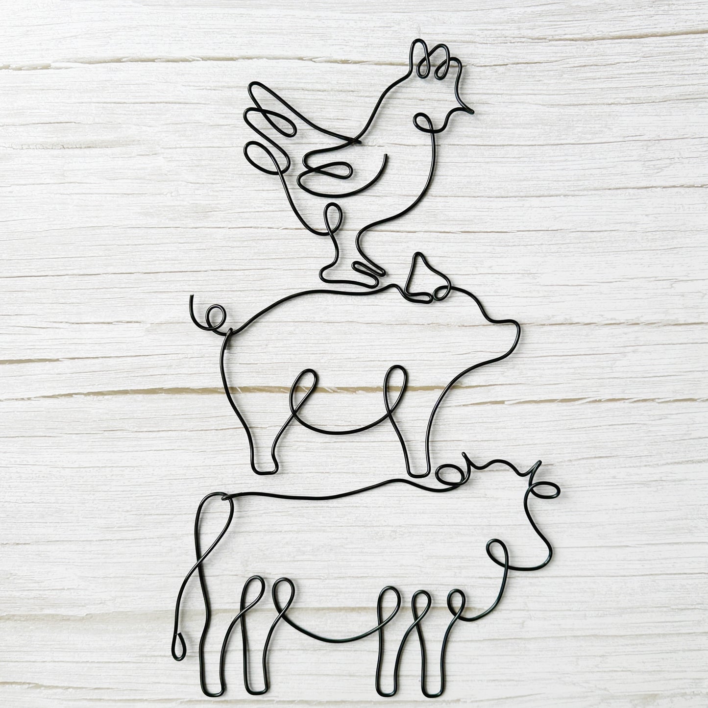 Black wire wall art featuring stacked farm animals: a chicken on a pig on a cow, styled against a rustic light wood background.