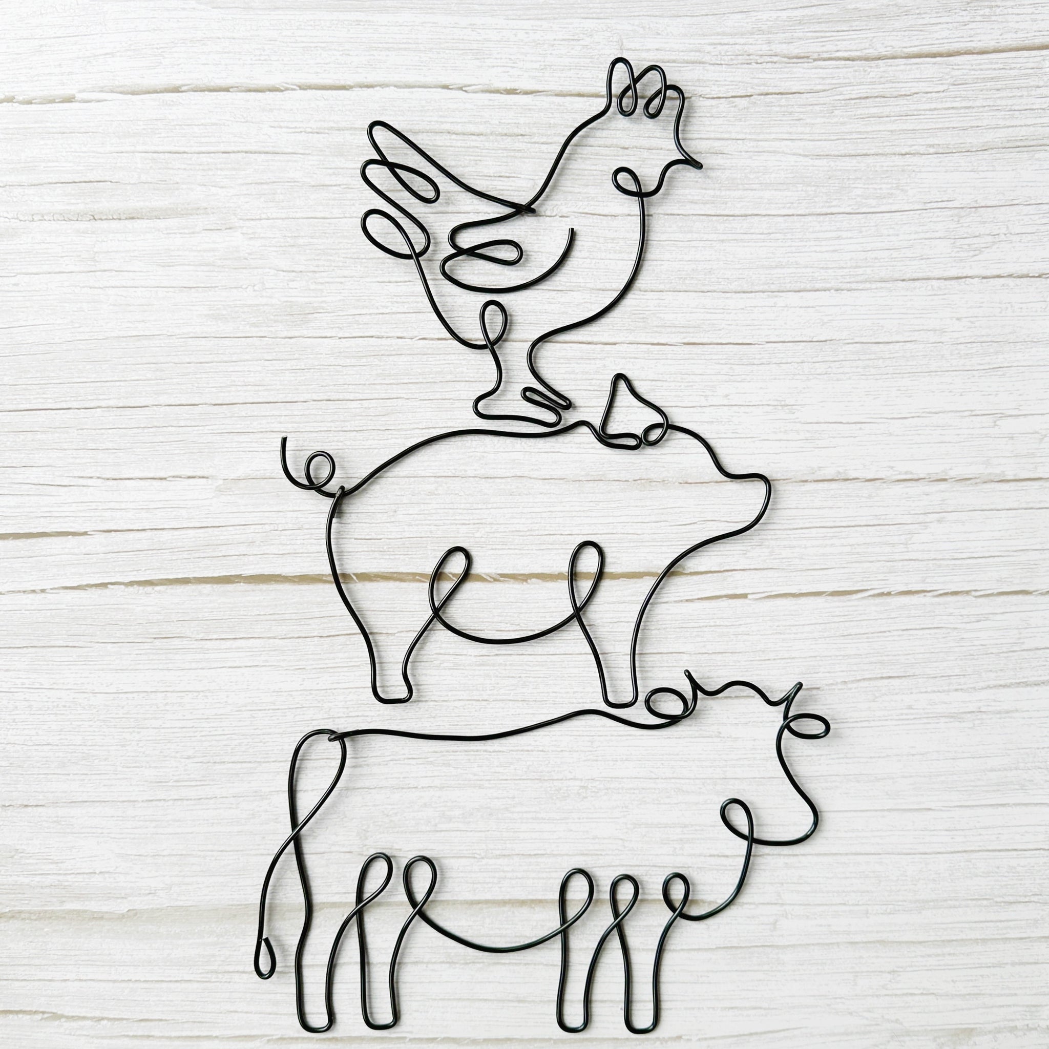 Black wire wall art featuring stacked farm animals: a chicken on a pig on a cow, styled against a rustic light wood background.