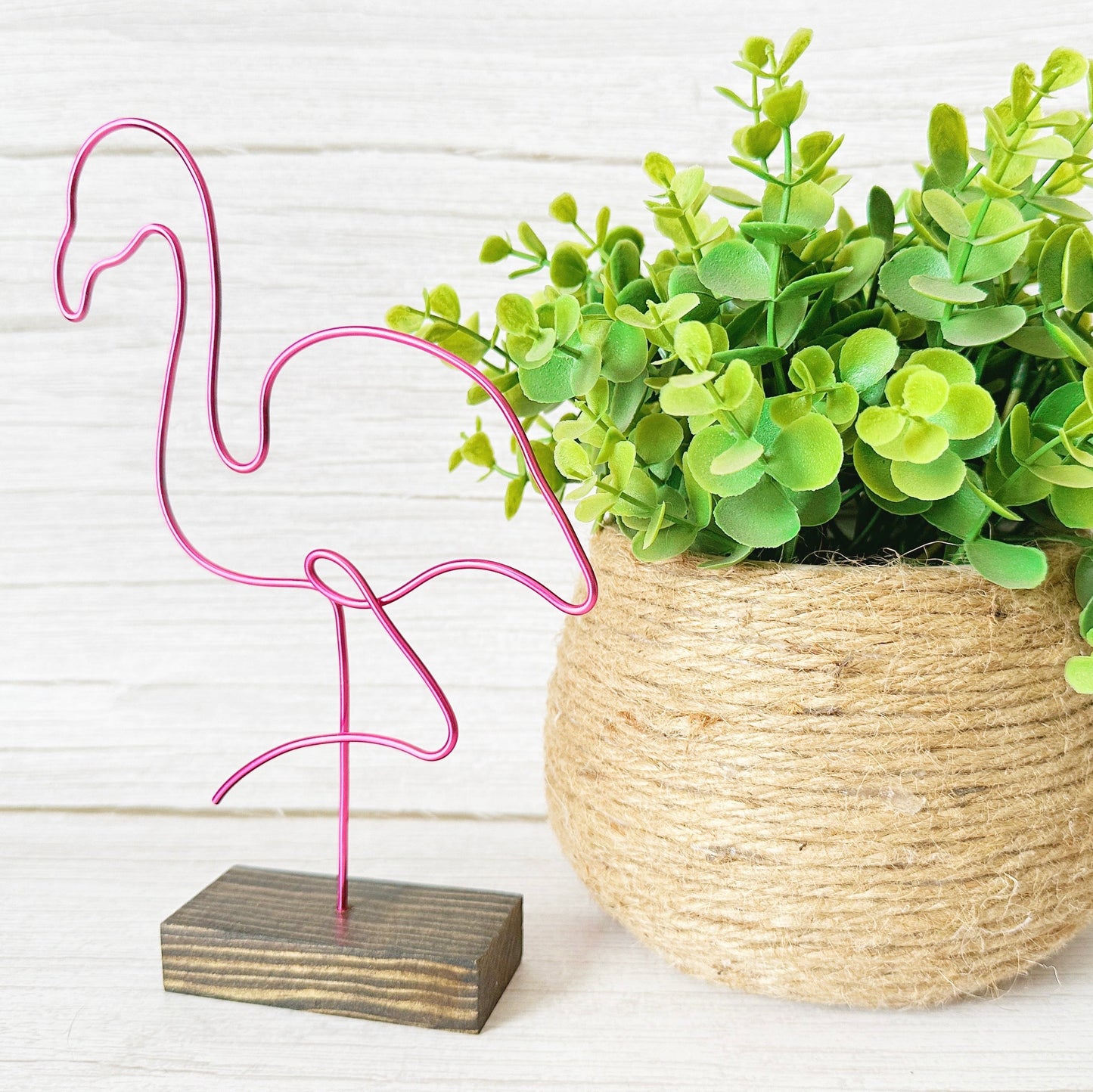 Pink flamingo decoration on a wooden base next to a potted plant with green leaves.